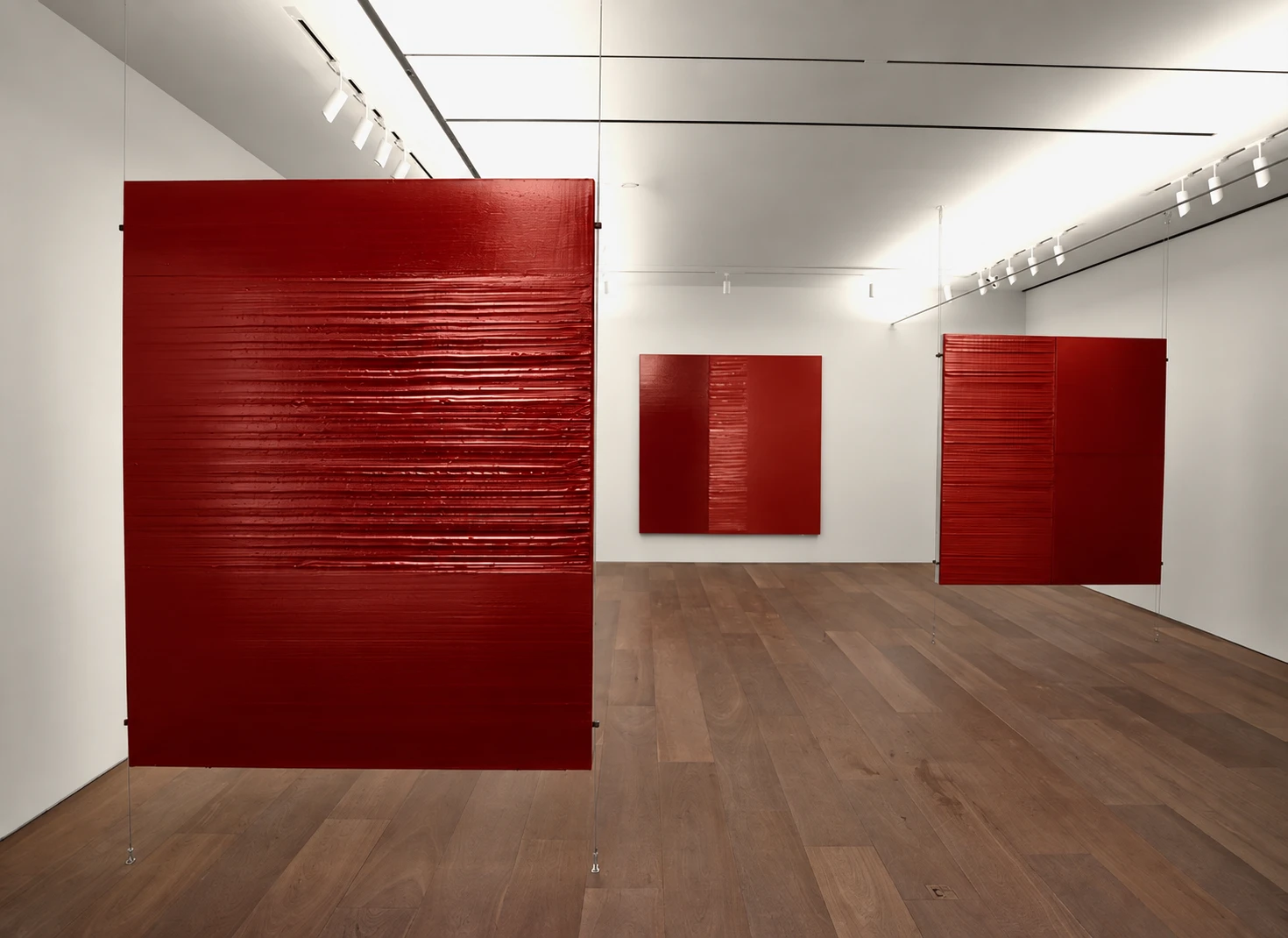 Three textured red panels, two hung on cables in a white gallery with a wooden floor.